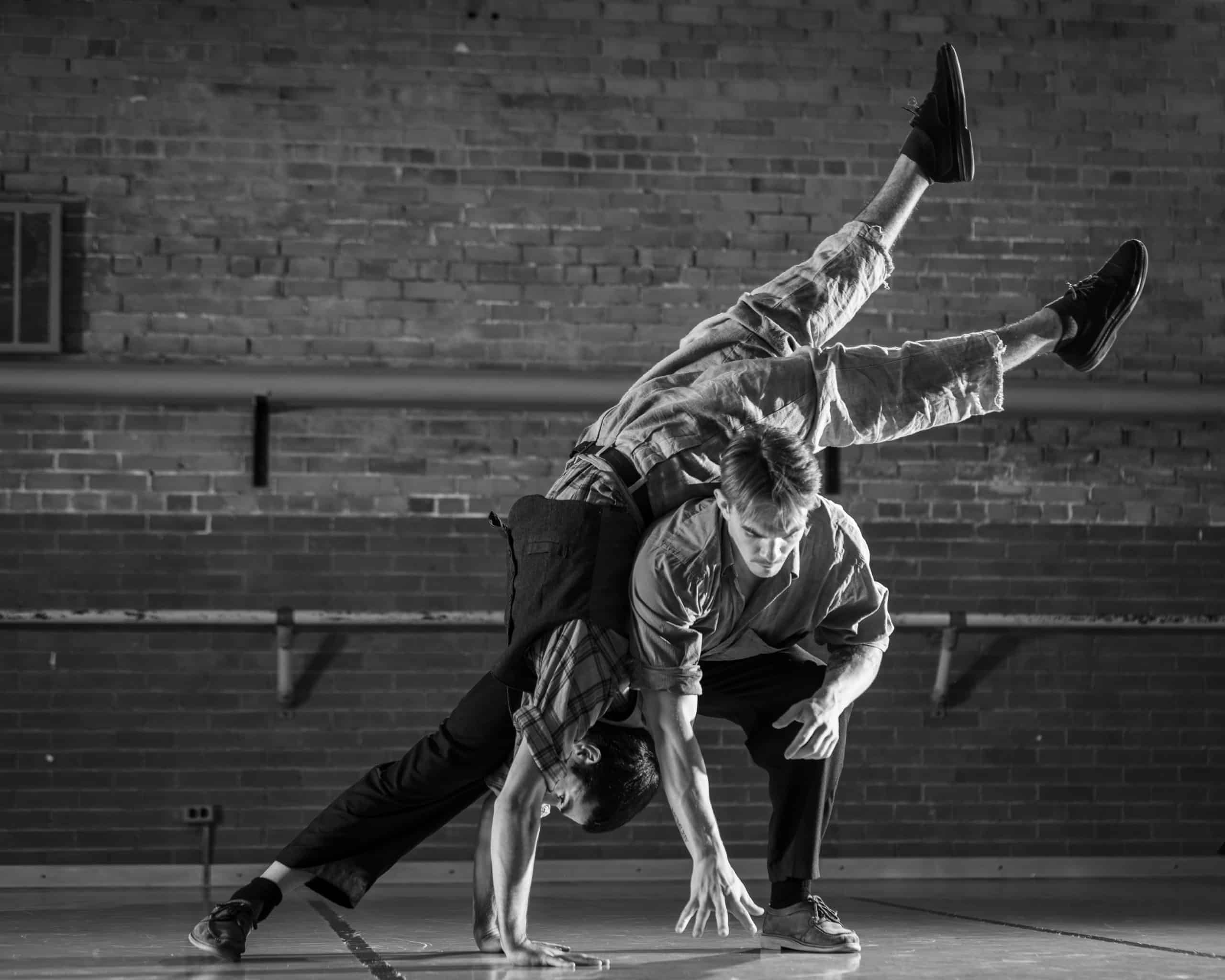 Black and white image of two men in studio with brick wall, one bent over supporting the other in a handstand going over his back.