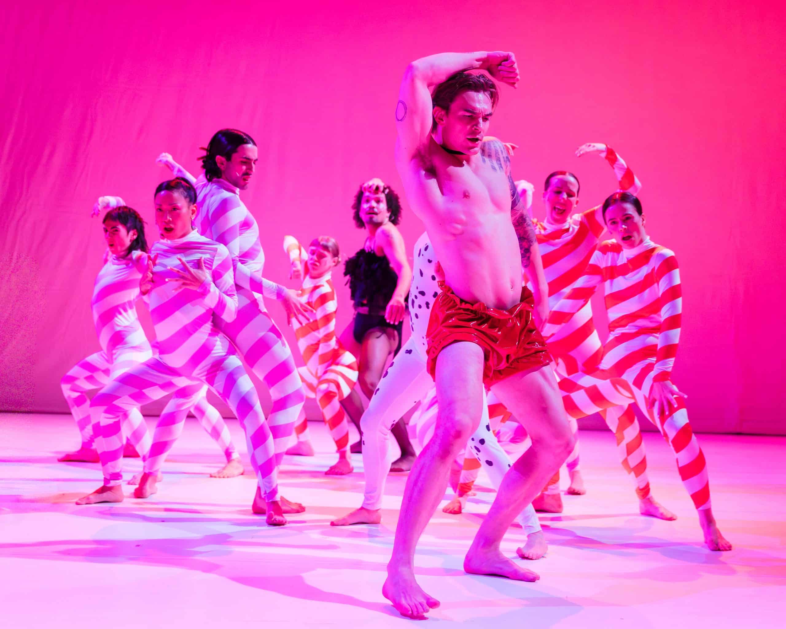 Dancers in candy cane unitards dancing around dancer in read shorts and bare torso.